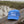 A light blue baseball cap with 'beach happy' embroidered in white on the front, resting on a wooden railing with a beach and dunes in the background.