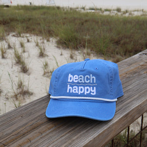 A light blue baseball cap with 'beach happy' embroidered in white on the front, resting on a wooden railing with a beach and dunes in the background.