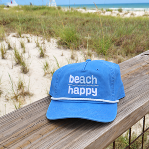 A blue baseball cap with 'beach happy' embroidered in white on the front, resting on a wooden railing with a beach and dune grass background.