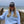 A woman standing on a wooden boardwalk by the beach, wearing a light blue sweatshirt and matching sweatpants with a black and white trucker hat that says 'BEACH HAPPY'.