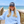 A smiling woman stands on a wooden boardwalk with the ocean and beach in the background, wearing a light blue hoodie and a navy trucker hat with white text that reads 'BEACH HAPPY'.