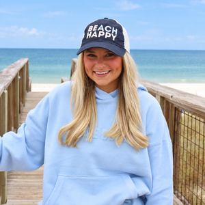 A smiling woman stands on a wooden boardwalk with the ocean and beach in the background, wearing a light blue hoodie and a navy trucker hat with white text that reads 'BEACH HAPPY'.