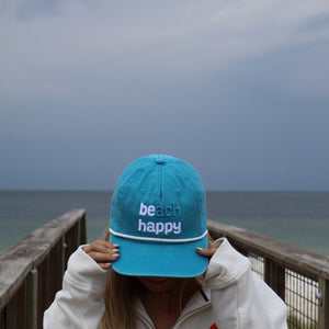 A woman wearing a white zip-up hoodie stands on a wooden boardwalk with the ocean and cloudy sky behind her, holding a bright turquoise baseball cap in front of her face. The cap has 'beach happy' embroidered in white on the front.
