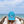 A person on a wooden boardwalk by the ocean wears a bright turquoise baseball cap with 'beach happy' embroidered in white on the front, holding the brim over their face.