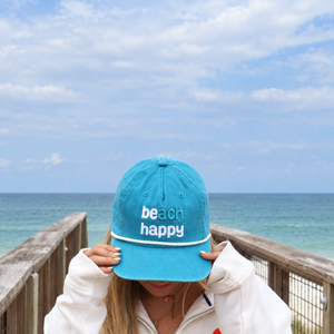 A person on a wooden boardwalk by the ocean wears a bright turquoise baseball cap with 'beach happy' embroidered in white on the front, holding the brim over their face.