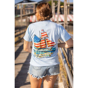 Person wearing a light blue t-shirt with a ship and American flag design, standing on a wooden dock.
