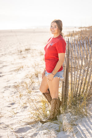Woman in a red shirt and denim shorts standing on a sandy beach with a wooden fence in the background.