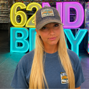 A woman with long blonde hair stands in a retail store wearing a blue t-shirt and a gray baseball cap with a '62nd Battalion' patch. Behind her are large neon signs reading '62ND BAY' in yellow, pink, and blue, with shelves of small items on a wooden wall above.