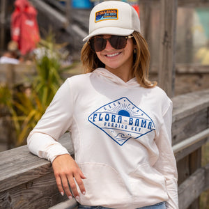 woman wearing a beige hoodie with the Flora-Bama text across the front with waves and sun and a white flora bama hat with colorful patch