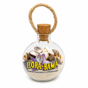 Glass bottle with cork lid containing seashells and sand, branded 'Flora-Bama'.