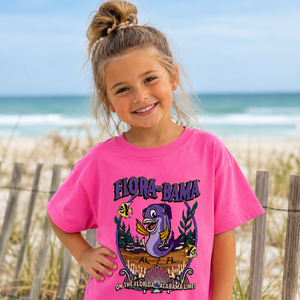 A young girl with her hair in a bun stands on a beach dune wearing a bright pink graphic t-shirt that reads 'FLORA-BAZA' with a purple seahorse and tropical design. The background shows the ocean, sky, and wooden fence.