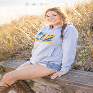 woman sitting on a boardwalk in light gray hoodie sweatshirt with beach scene and flora bama across the front center