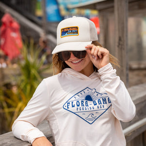 A person wearing a white baseball cap with blue and orange embroidery on the front panel, featuring the text 'Flora-Bama GET PERDIDO 1964' and cream flora bama long sleeve hoodie with blue flora bama text on the front