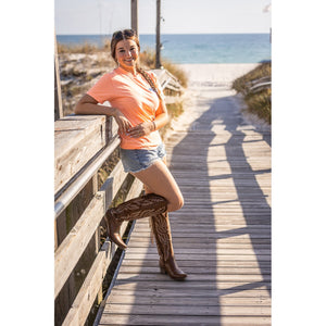Woman in peach shirt and denim shorts with cowboy boots standing on a wooden boardwalk by the beach.