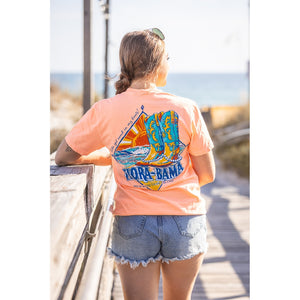 Person wearing a peach t-shirt with a graphic design, standing on a wooden boardwalk by the beach.