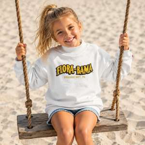 A young girl with blonde hair in a ponytail sits on a wooden swing on a sandy beach, wearing a white long-sleeve sweatshirt with a yellow and black 'FLORA-BAMA' graphic on the chest.