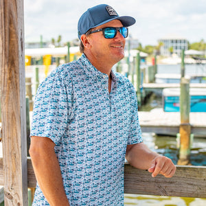 Man wearing a blue patterned shirt, cap, and sunglasses by a dock with boats in the background.