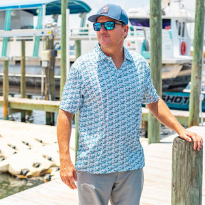 Man standing on a dock wearing a blue patterned shirt and sunglasses, with boats in the background.