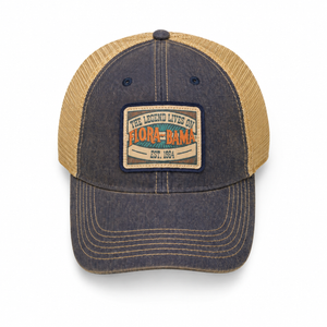 A trucker-style baseball cap with a dark gray washed denim front and brim, and tan mesh back panels. It features a rectangular embroidered patch on the front with the text 'THE LEGEND LIVES ON FLORA AND GAMA EST.