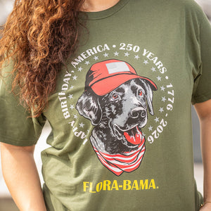 Person wearing a green t-shirt with a graphic of a dog wearing a red cap and bandana, celebrating America's 250th birthday.
