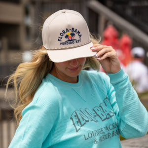 Person wearing a light blue sweatshirt with branding and a beige cap with flora bama text, standing outdoors.