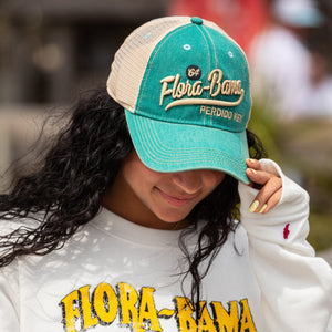 Person wearing a teal hat with mesh background and flora bama script embroidered text on the front