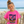 A young girl with her hair in a bun stands on a beach dune wearing a bright pink graphic t-shirt that reads 'FLORA-BAZA' with a purple seahorse and tropical design. The background shows the ocean, sky, and wooden fence.