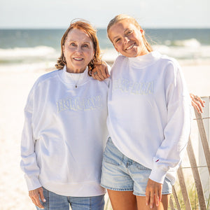 Two people wearing white sweatshirts with flora bama text, standing on a beach.