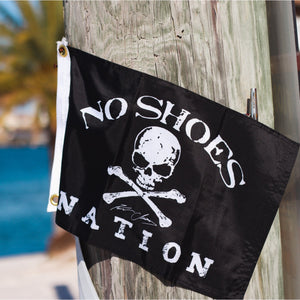 Black flag with 'No Shoes Nation' text and skull design hanging on a wooden post by a poolside.