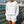 A woman wearing a white crewneck sweatshirt and matching shorts with 'FLORA-BAMA' text, standing on a wooden boardwalk with a beach and ocean in the background.