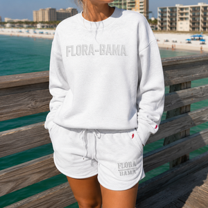A woman wearing a white crewneck sweatshirt and matching shorts with 'FLORA-BAMA' text, standing on a wooden boardwalk with a beach and ocean in the background.