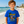 A young boy stands on a sandy beach wearing a bright blue graphic t-shirt with a 'Flora-Bama' cartoon design and khaki shorts. The background shows the ocean, pier, and beach houses.