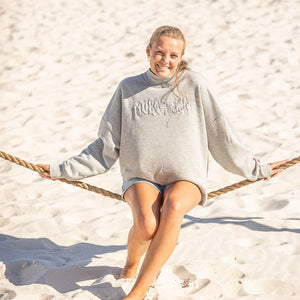 Woman sitting on a rope swing on a sandy beach with gray sweatshirt with flora bama text