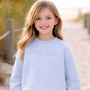 Young girl wearing a light blue sweatshirt with 'FLORA-BAMA' text, on the front